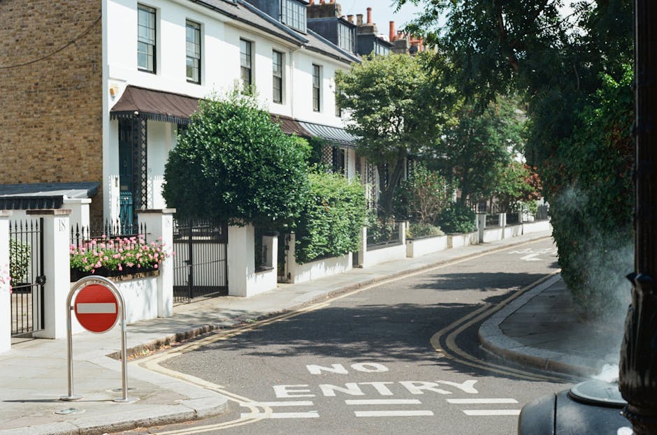 A quiet residential street scene showing white terraced houses with dark-colored awnings over the entrances, surrounded by well-maintained green shrubs and trees. The pavement is lined with flower bed