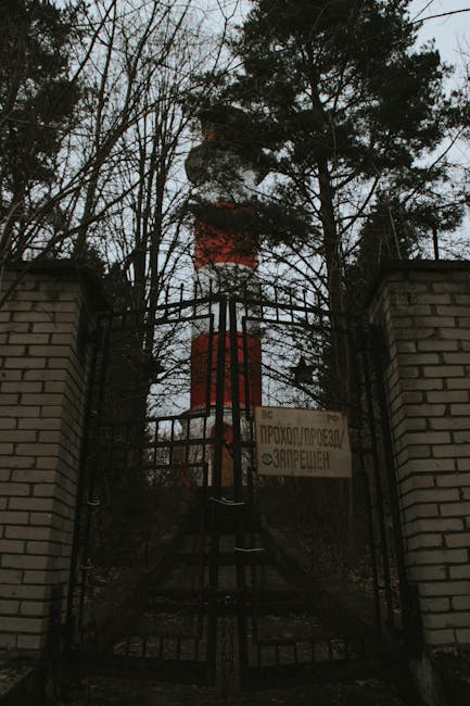 A metal gate between two brick pillars leads to a property with a view of a tall red and white smokestack in the background, partially obscured by leafless trees. The gate appears closed and is secured with a latch, with a sign attached to it displaying text in Cyrillic script, indicating restricted access. The surrounding environment is outdoors, with overcast lighting conditions, and the area appears to be part of an industrial or utility site. No moving or loading activities are visible; the image focuses on the gate, the signage, and the distant smokestack, offering a neutral, factual depiction of the scene as part of an industrial or urban landscape.
