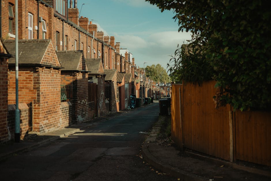 A narrow residential street in Forest Gate lined with brick terraced houses featuring small front gardens, some with brickwork walls and sloped tiled roofs over doorways. The street is paved with asphalt and has several waste bins placed along the curb, indicating waste collection or clearance operations. On the right side of the image, a tall wooden fence separates the pavement from a garden or backyard. The scene appears to be during late afternoon or early evening with soft, natural lighting and partly cloudy sky overhead. Several trash bags and cardboard boxes are visible near the waste bins, suggesting clearance of bulky waste items as part of a home relocation or moving process. This context aligns with the services provided by Man with Van Forest Gate, who handle removals and furniture transport in the area, involving loading and moving activities typical of household shifts on terraced streets.