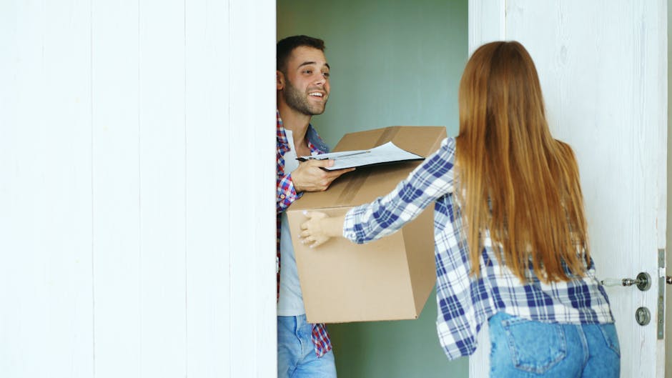 A man with dark hair, a beard, and a cheerful expression stands inside a doorway, receiving a large cardboard box from a woman with long red hair, wearing a blue and white checked shirt and jeans. The woman is handing the box to the man, who is holding some papers or a clipboard. The background shows a light green wall and part of a white door frame. The scene depicts a home relocation process, with the woman assisting in moving boxes and household items. This interaction occurs in an indoor setting, with the individuals engaged in loading or unloading furniture and packing materials, including cardboard boxes, during a house move arranged by Man with Van Forest Gate, a professional removals service covering estate flats and relocation logistics.