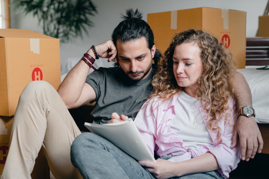 A young couple, a man with dark hair tied in a bun and a woman with curly blonde hair, sit on the floor amidst moving boxes during a house relocation in Forest Gate. The man wears a grey t-shirt and beige trousers, leaning against a cardboard box with his arm around the woman, who is dressed in a light pink shirt and grey jeans. They are reviewing a document or packing list together, with focused expressions on their faces. The background shows several large cardboard boxes sealed with packing tape, some marked with red symbols, and a bed with pillows partially visible. The scene is indoors, with natural light illuminating the space, capturing the typical packing and moving process involved in a home relocation, as undertaken by services such as Man with Van Forest Gate.