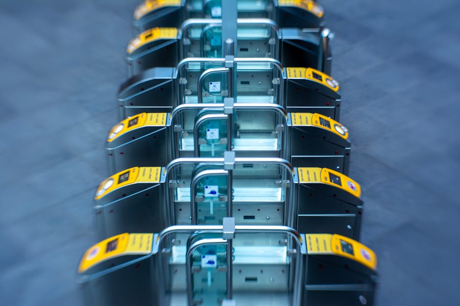 Inside a busy train station, metal and glass ticket barriers with illuminated green lights are arranged in a row across the foreground. Behind the barriers, several passengers are visible, including a