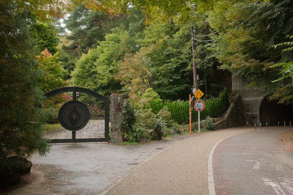 A purple scooter parked on a gravel driveway in front of a decorative metal garden archway with intricate scrollwork, leading into a lush, green wooded area with tall trees and dense foliage. The scoo