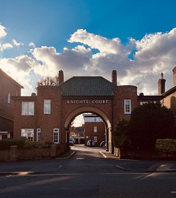 The image shows a brick building with an arched entryway labeled 'Knights Court,' located within a residential area. The building features two square towers on either side of the arch, with small windows and a gray tiled roof. Behind the arch, a street with parked cars and additional brick houses is visible, indicating an urban or suburban neighbourhood. The sky above is partly cloudy with a mix of white clouds and blue sky, and sunlight casts shadows on the building and pavement. When coordinating house removals or furniture transport, companies like [COMPANY_NAME] may operate in such areas, with careful loading and unloading taking place outside these types of residential buildings, often involving the use of trolleys, blankets, and moving straps to ensure safe equipment handling during home relocation or packing and moving processes.
