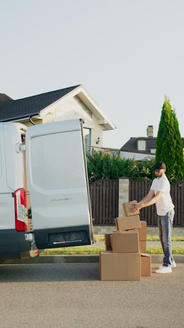 A man wearing a black cap, black vest, and blue long-sleeve shirt is outside a house, pushing a hand truck loaded with two large cardboard boxes wrapped with plastic straps. He is standing on a paved 